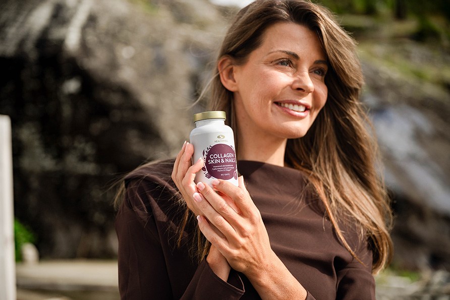 Leende kvinna med långt brunt hår och brun tröja håller upp en burk Healthwell Collagen Skin & Nails utomhus framför en klippa.