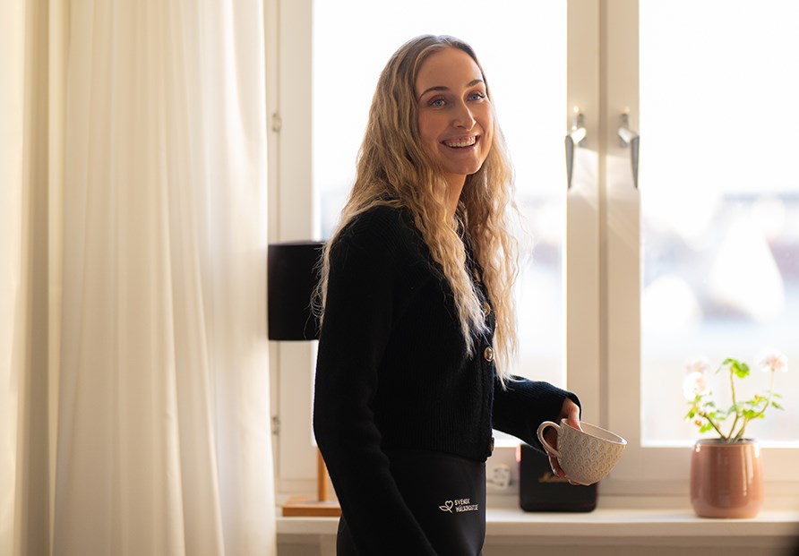 Annie vid fnstret med en kopp i handen.