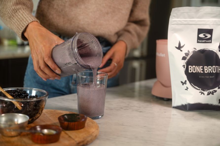En kvinna hller upp blbrssmoothie med benbuljong frn en mixer i ett glad. P bordet syns en skl med blbr och en pse Healthwell Benbuljong EKO.