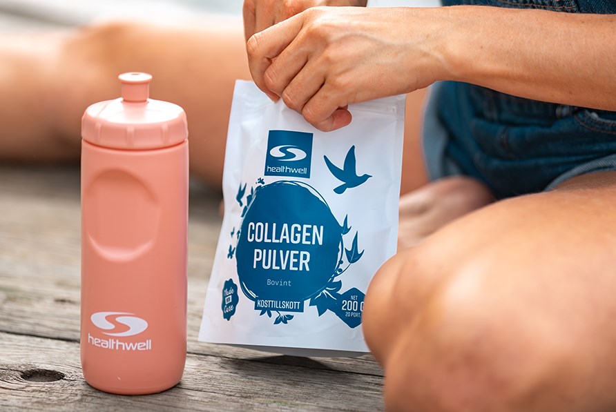 Healthwell Collagen Pulver Bovint 2.0 visas i nrbild p en brun brygga. ven en vattenflaska frn Healthwell r synlig, samt en kvinnas ben, men dessa r inte i fokus. 