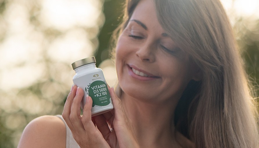 Glad kvinna håller i Healthwell Vitamin D3 5000 + K2 100 ute i naturen.