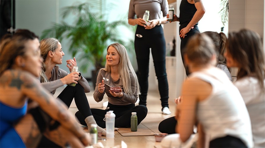 En grupp kvinnor sitter tillsammans p yogamattor i ett ljust rum och pratar medan de ter och dricker hlsosamma mellanml och juicer.