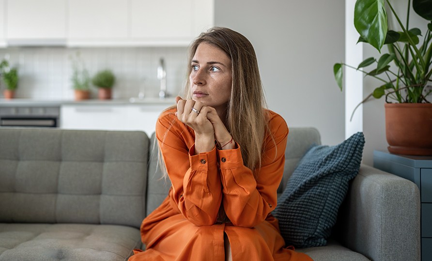 En kvinna sitter p� en soffa med huvudet i h�nderna och tittar ut i luften.