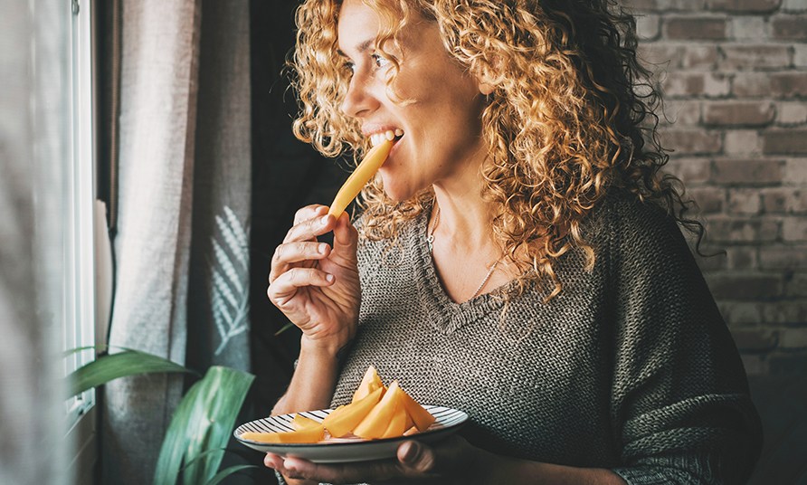 En kvinna st�r vid ett f�nster och �ter en bit frukt.