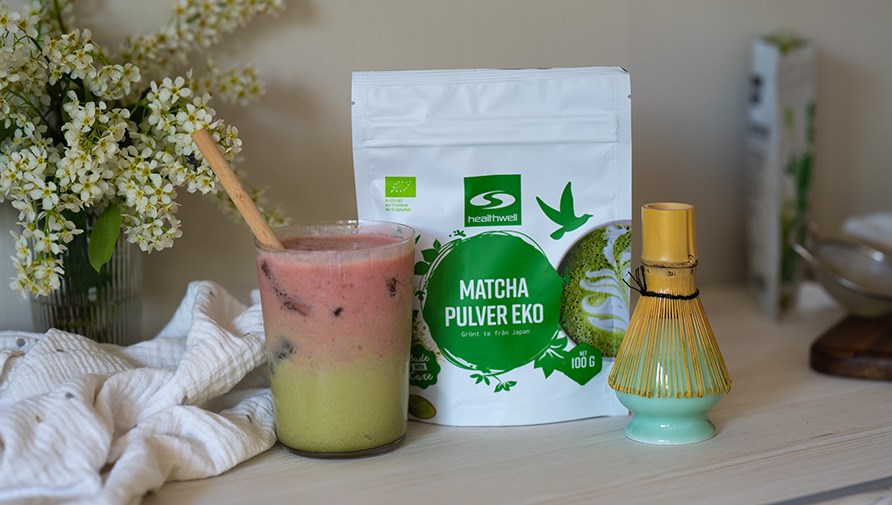 Healthwell Matcha Pulver EKO i originalf�rpackning, iced strawberry matcha latte i ett glas med ett bambusugr�r, visp f�r matchalatte, blombukett i en vas och en k�kshandduk p� ett bord. 